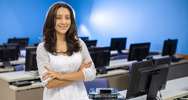 Young woman smiles with arms crossed, standing in a computer lab at 澳门线上博彩.