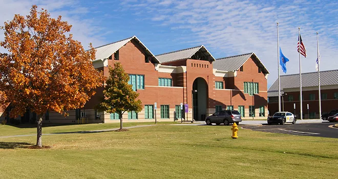 Entrance of 澳门线上博彩 West Campus. Tree with orange leaves in the foreground. Oklahoma and American flags fly on flag poles.